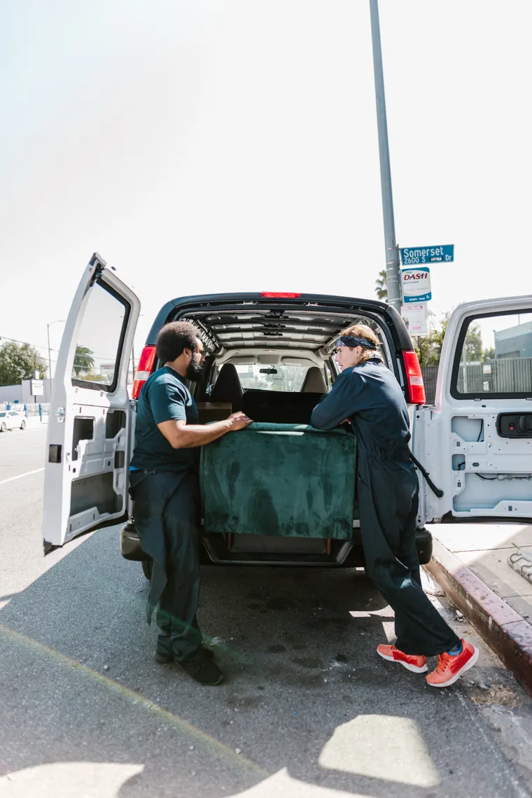 Two-person crew loading an old sofa into the back of a hauling van at the curb